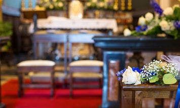 Bouquet of white and blue flowers sits on a wooden pew in the foreground, with an altar and two chairs covered in white fabric in a decorated church setting.