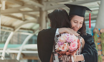 A graduate in a cap and gown hugs someone while holding a bouquet of flowers, standing in an open, modern architectural space with glass and metal structures.