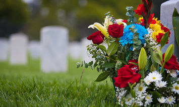 A bouquet of colorful flowers, including red roses and lilies, rests on a grassy area near gray tombstones in a cemetery.