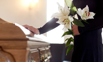 A person in black clothing holds white lilies and rests a hand on a wooden casket in a softly lit room, suggesting a funeral setting.