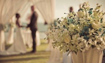 A white flower arrangement sits in the foreground while a couple exchanges vows under a white fabric-draped arch in the blurred background of an outdoor setting.